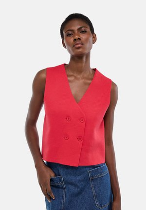 Femme portant un gilet rouge sans manches à double boutonnage et une jupe en jean bleue avec de grandes poches avant, main posée casual dans la poche, fond neutre.