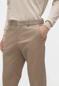 Personne portant un pantalon décontracté beige avec la main dans la poche droite et un haut à manches longues beige clair sur un fond uni.