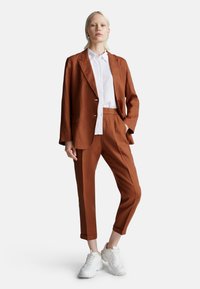 Liniový blazer a kalhoty v rzi barvě, doplněné bílou košilí s knoflíky a bílými teniskami. Blazer má volný střih a klopové límce.