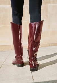 Lśniące bordowe botki do kolan z bocznym zamkiem, z łańcuchowym akcentem w okolicy kostki i płaską czarną podeszwą. Gładka, błyszcząca tekstura.
