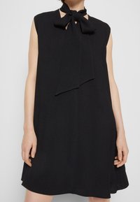Svart ärmlös klänning med knytning vid halsen, lätt tyg, lös passform och subtil strukturerad yta. Inga synliga mönster eller hårdvara.