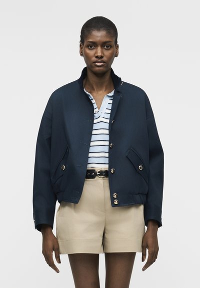 Femme portant une veste marine boutonnée par-dessus un polo rayé bleu et blanc, un short beige taille haute et une ceinture noire.