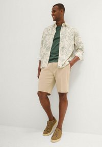 Outfit casual composto da una maglietta a maniche corte beige chiaro con motivo a foglie verdi, una maglietta verde, pantaloni corti beige e sneakers color sabbia.