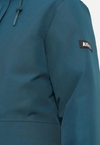 Casaco exterior em teal com acabamento mate, apresentando um capuz ajustável e um rótulo preto no punho. Material impermeável.