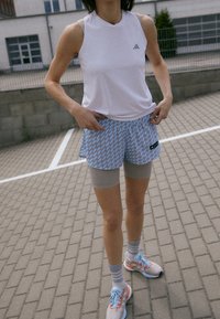 Osoba oblečená v bílém bezrukávovém topu Adidas, modrých vzorovaných šortkách, šedých cyklistických šortkách pod nimi a běžeckých botách stojící na chodníku.