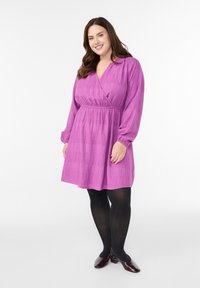 Violeta ietīta kleita ar gariem pūkainiem piedurknēm, teksturētu audumu, elastīgu vidukļa joslu un izplešamu svārku, kopā ar melnām zeķbiksēm un bordo krāsas augstpapēžu kurpēm.