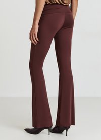 Bordeauxfarbene, ausgestellte Hose aus einem glatten Stretchmaterial mit hohem Bund. Sie zeigt eine einheitliche Farbe mit minimalen Details. Kombiniert mit schwarzen High Heels.