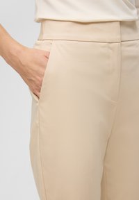 Pantalons beiges en tissu lisse et texturé, avec une poche latérale et une taille structurée, montrés de la taille jusqu'à la main.