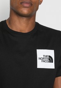 Juodas medvilnės marškinėliai su balta kvadratine krūtinės etikete, ant kurios juodu tekstu ir logotipu „THE NORTH FACE“. Klasikinio apvalaus kaklo dizainas.