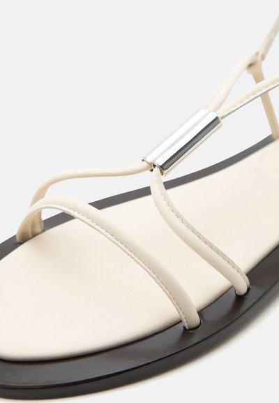 Μπεζ σανδάλι με λεπτούς ιμάντες και ασημένια κυλινδρική αγκράφα σε μαύρη σόλα, που δείχνει την περιοχή των μπροστινών δακτύλων των ποδιών.