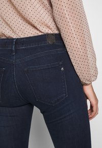 Dark blue denim jeans with a fitted design and subtle stitching details. Features back pockets and a leather logo patch at the waist.