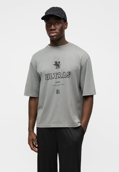 Sivá bavlnená tričko s krátkymi rukávmi, ozdobené čiernym nápisom "ULTRAS" a grafikou leva. Kombinované s čiernymi nohavicami a jednoduchou čiapkou.
