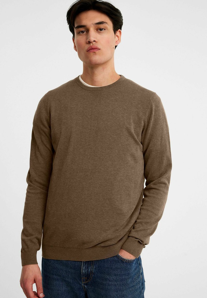 Bruine gebreide trui met een ronde halslijn en lange mouwen, met ribgebreide manchetten en zoom, gedragen over een witte ondershirt.