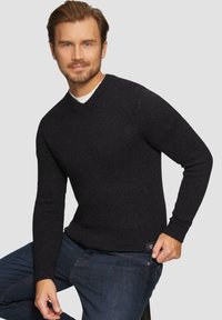 Schwarzer gestrickter Pullover mit V-Ausschnitt und langen Ärmeln, der ein strukturiertes Muster und ein kleines Etikett an der Saumkante aufweist, kombiniert mit dunklen Jeans.