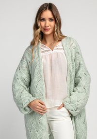 Jasnozielony, dziergany kardigan z teksturowanym, ażurowym wzorem. Modelka ma na sobie białą bluzkę, co dopełnia warstwowy wygląd.