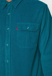 Camicia in corduroy verde acqua con colletto button-down, caratterizzata da una singola tasca sul petto e pulsanti blu coordinati. Leggera texture a coste su tutta la superficie.
