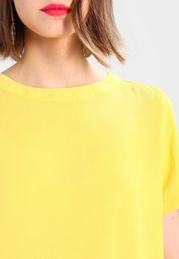 Žlutá blůzka s krátkým rukávem a kulatým výstřihem, vyrobená z lehkého materiálu, s hladkou texturou a čistými liniemi.