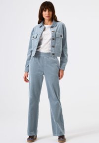 Femme portant une veste et un pantalon en velours côtelé bleu clair avec un top en dentelle blanche et des baskets marron, se tenant devant un fond blanc uni.