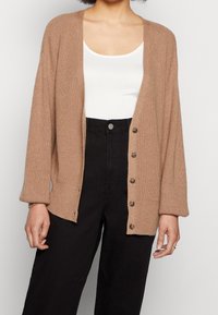 Frau trägt eine beigefarbene gestrickte Cardigan mit Schildkrötenmuster-Knöpfen, ein weißes Oberteil mit Rundhalsausschnitt und eine hoch taillierte schwarze Hose, die vor einer weißen Wand steht.