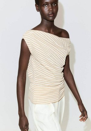 Donna dalla pelle scura che indossa un top asimmetrico senza maniche a righe beige e bianco e pantaloni bianchi, in piedi contro uno sfondo chiaro e semplice.
