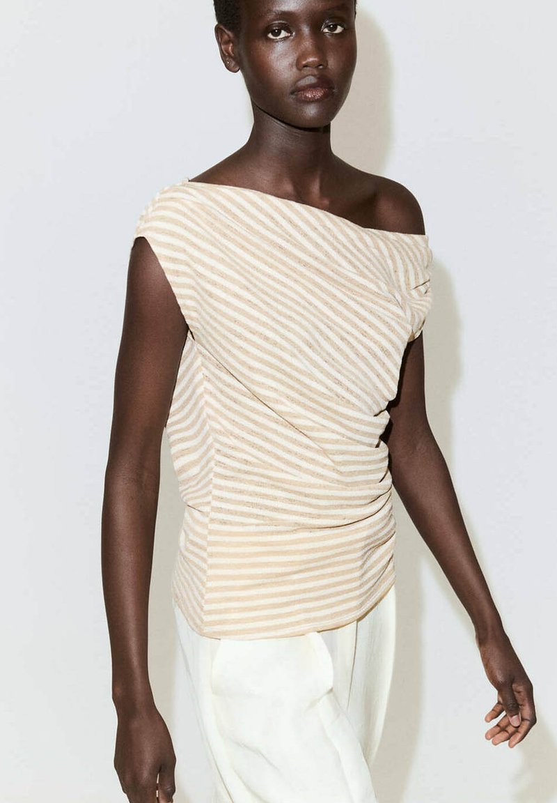 Femme à la peau foncée portant un haut asymétrique sans manches beige et blanc à rayures et un pantalon blanc, debout devant un fond clair uni.