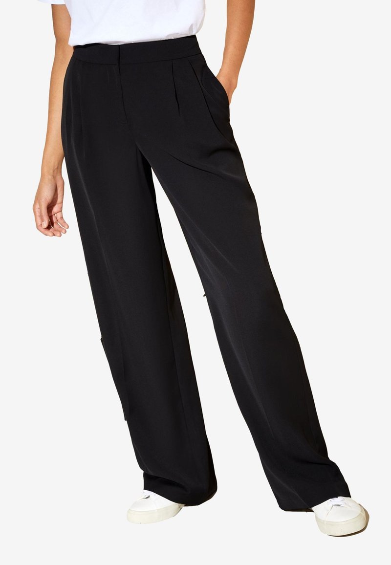 Mujer con pantalones negros de pierna ancha con bolsillos laterales, combinados con zapatillas blancas y una camiseta blanca, de pie con una mano en el bolsillo.