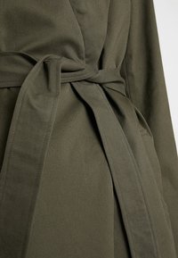 Manteau en tissu doux olive, ceinturé avec une ceinture nouée, présentant une texture lisse et des lignes épurées. Comprend des poches latérales et un col classique.