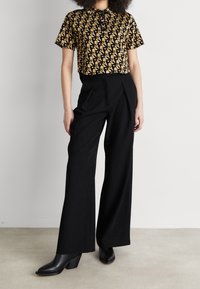 Femme portant un haut court à motif géométrique beige et noir, un pantalon large taille haute noir et des bottes à talons noires.