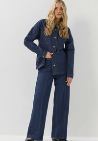 Ensemble en jean, composé d'une veste et d'un pantalon à jambes larges, bleu marine, avec des boutons dorés et des poches, mettant en avant des coutures contrastées et une coupe décontractée.