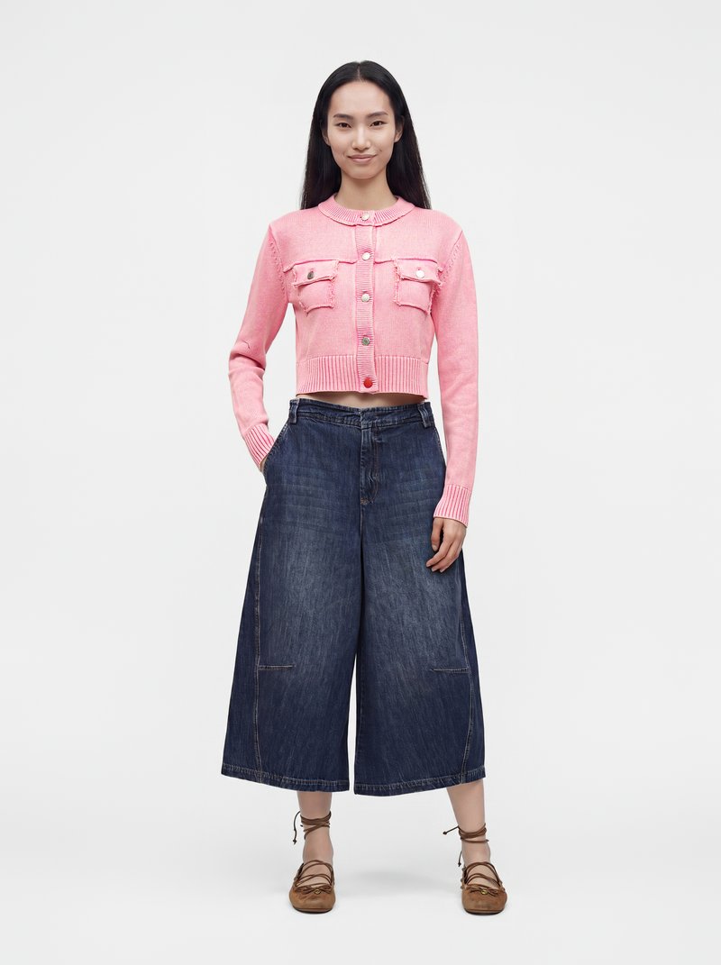 Femme debout portant un cardigan court rose boutonné, un pantalon large en denim bleu foncé et des chaussures plates marron à lacets sur fond blanc.