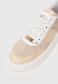 Sneaker beige e bianco con lacci bianchi in satin e tag dorato "Steve Madden" vicino alla punta su uno sfondo bianco.