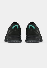 Sapatos de desporto pretos com uma superfície texturizada, apresentando um detalhe em verde menta e sola de borracha, mostrando a vista traseira com um padrão de piso visível.