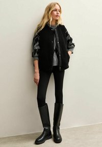 Svart ärmlös väst med fluffig textur över en grå argyle-tröja, i kombination med tajta svarta leggings och knähöga svarta stövlar.