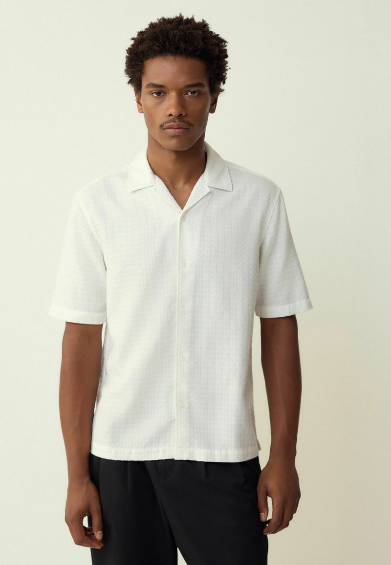 Short-sleeve white shirt with a textured pattern, button closure, and a fold-down collar, paired with black trousers.