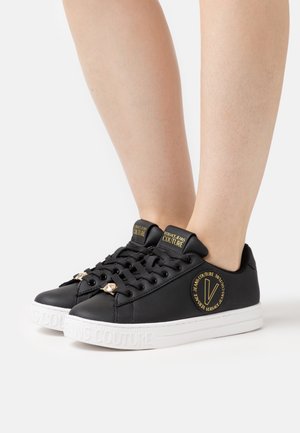 Zwarte leren sneakers met gouden accenten en branding op de tong en zijkant, voorzien van platte veters en een gestructureerde witte rubberen zool.