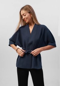 Marinblå kimonotopp med korta vida ärmar, knytbälte i midjan och slät textur. Svarta byxor kompletterar outfiten.