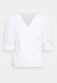 Blouse cache-cœur blanche avec manches ballon, tissu texturé avec de petits points en relief et décolleté plongeant en V. Détail à cordon de serrage sur le côté.