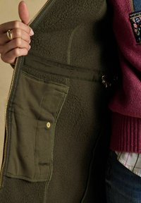 Ręka trzyma otwartą zieloną kurtkę z polaru, pokazującą wewnętrzną kieszeń z zatrzaskiem i złotym zamkiem błyskawicznym wzdłuż krawędzi.