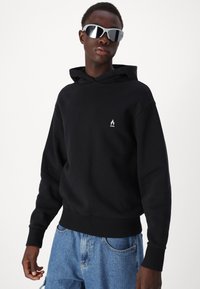 Hoodie en coton noir avec manches longues, une poche avant et un petit logo de flamme sur la poitrine gauche. Porté avec un short en denim bleu clair.