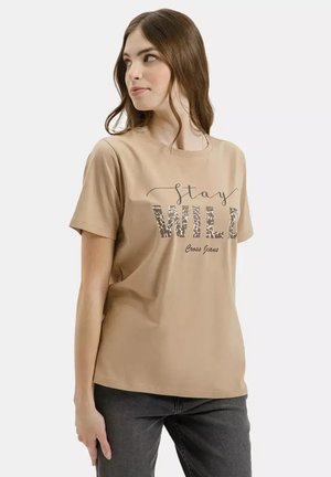 Kobieta w beżowej koszulce z napisem "Stay WILD Cross Jeans" w panterkowy wzór, w ciemnoszarych dżinsach, patrząca w bok.