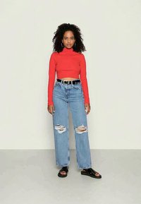 Top cropped de mangas longas vermelho combinado com jeans de perna larga azul claro com detalhes rasgados. Cintos pretos e sandálias pretas de ponta aberta completam o visual.