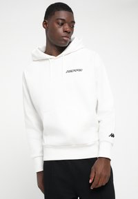 Junger Mann in einem weißen Kappa-Hoodie mit Fronttasche und schwarzen Hosen, der leicht nach unten und nach links schaut.