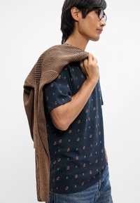 Ναυτικό κοντομάνικο μπλουζάκι με πολύχρωμα λουλουδάτα σχέδια, συνδυασμένο με καστανή ρίγανη ζακέτα ριγέ ριγμένο στον έναν ώμο, φορεμένο με μπλε τζιν.