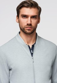 Giovane uomo con capelli castano chiaro e barba, che indossa una giacca grigia chiara con zip su una maglietta scura, che guarda direttamente davanti a sé su uno sfondo semplice.