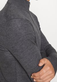 Grå åtsittande tröja med hög krage, långa ärmar och en texturerad stickad mönster. Har en halv dragkedjestängning vid nacken.