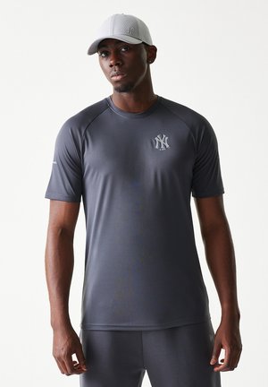 Mand iført mørkegrå New York Yankees sportstrøje og matchende lysegrå baseballkasket, stående mod en ensfarvet baggrund.