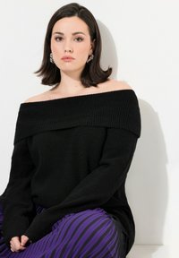 Schwarzer Strickpullover mit Off-Shoulder-Ausschnitt und Rippstruktur, kombiniert mit einem lila-schwarz gestreiften Rock. Modell sitzt vor einem weißen Hintergrund.