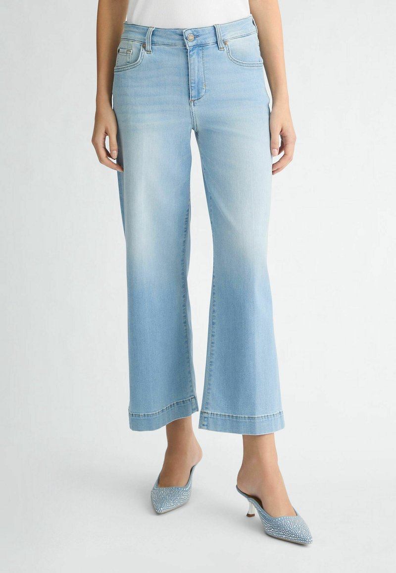 Mujer que lleva unos jeans de corte acampanado de tiro alto en azul claro y unos tacones de mule azul con puntera puntiaguda y adornos de strass, de pie sobre un suelo blanco.