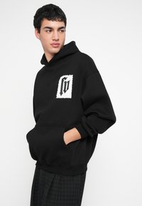 Čierna mikina s kapucňou a predným vreckom na brušku, s textúrovanou bielou nášivkou loga na ľavej strane. Mäkká látka, voľnejší strih.