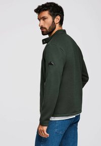 Mörkgrön, texturerad jacka med krage, sidofickor och en svart triangulär logo på vänster axel. Kombinerad med blå jeans.